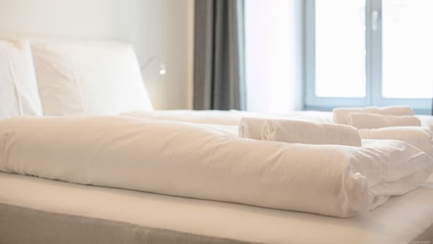 Una cama bien hecha con ropa blanca y almohadas, una ventana con cortinas, y una habitación luminosa.
