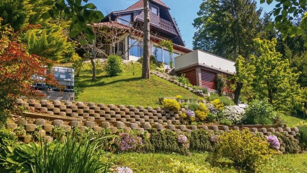A house with a sloped roof, a large glass window, and a garden with various plants and a stone wall. - Forest Retreat Bonidesa - Terrace & Jacuzzi (Ljubno)