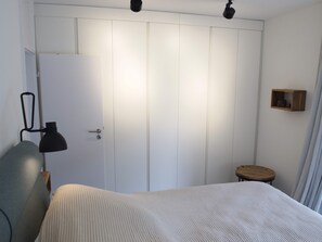 A bedroom with a bed, a wardrobe, a desk lamp, a small wooden stool, and a wall-mounted shelf.