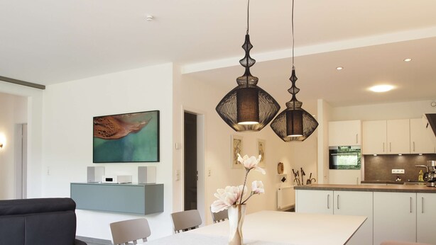 A modern dining area with a white table, black and grey chairs, and a vase with flowers. There is a kitchen in the background with white cabinets and a microwave.