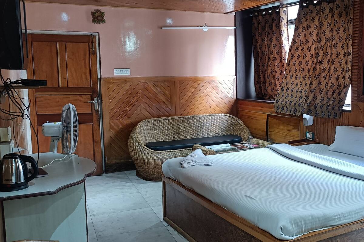 A room with a bed, a chair, a desk with a kettle, a fan, and curtains.