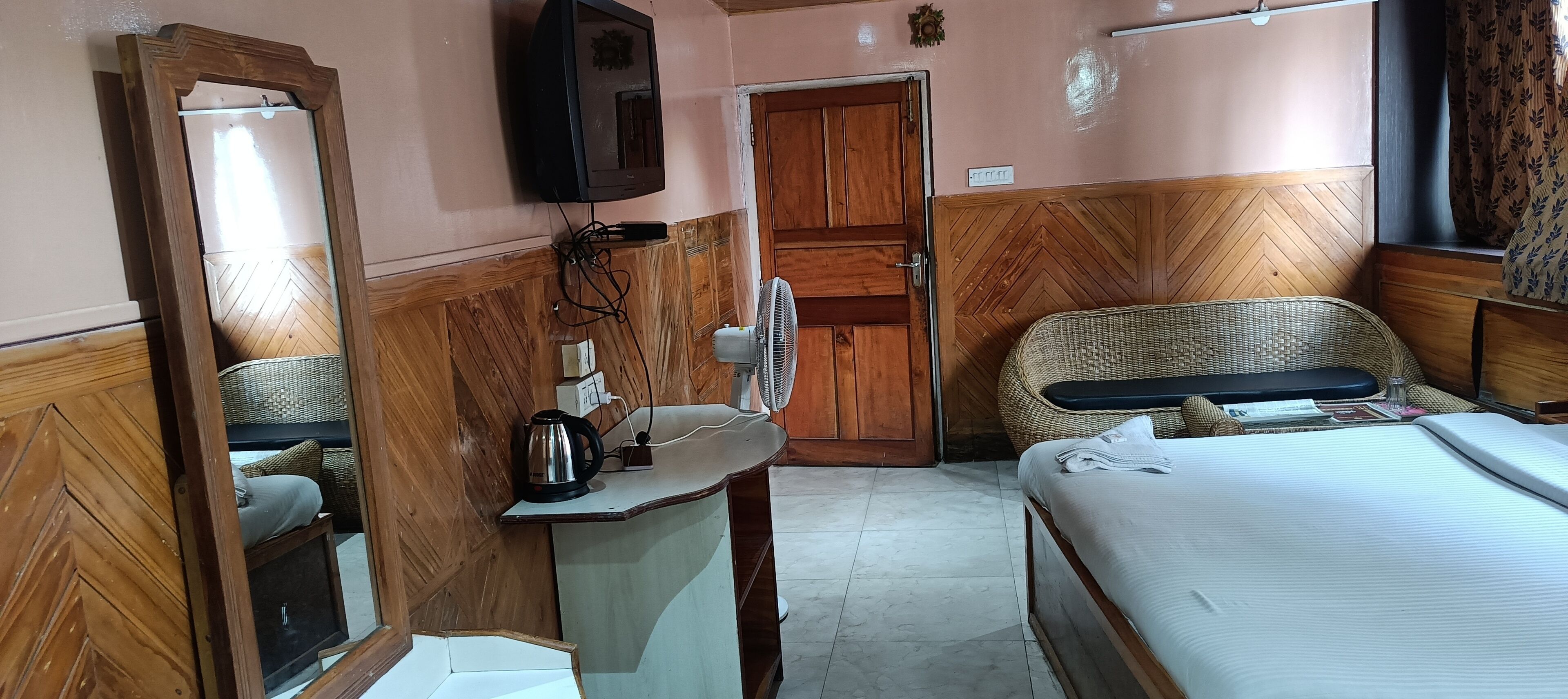 A compact hotel room with a bed, a small table with a kettle, a fan, and a television mounted on the wall.