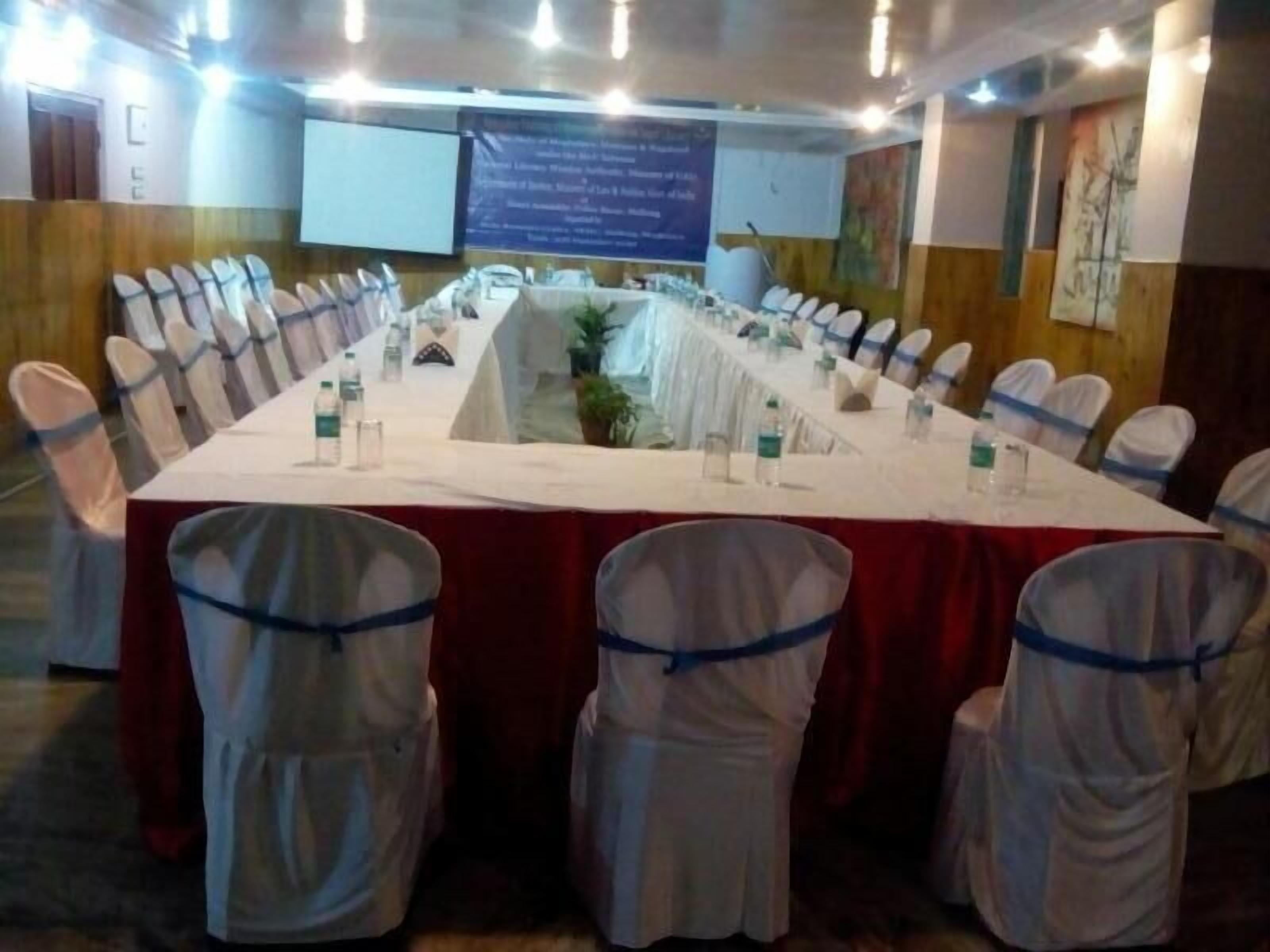 Un salón de conferencias con una larga mesa, sillas blancas con cintas azules y una pantalla de proyector al fondo.