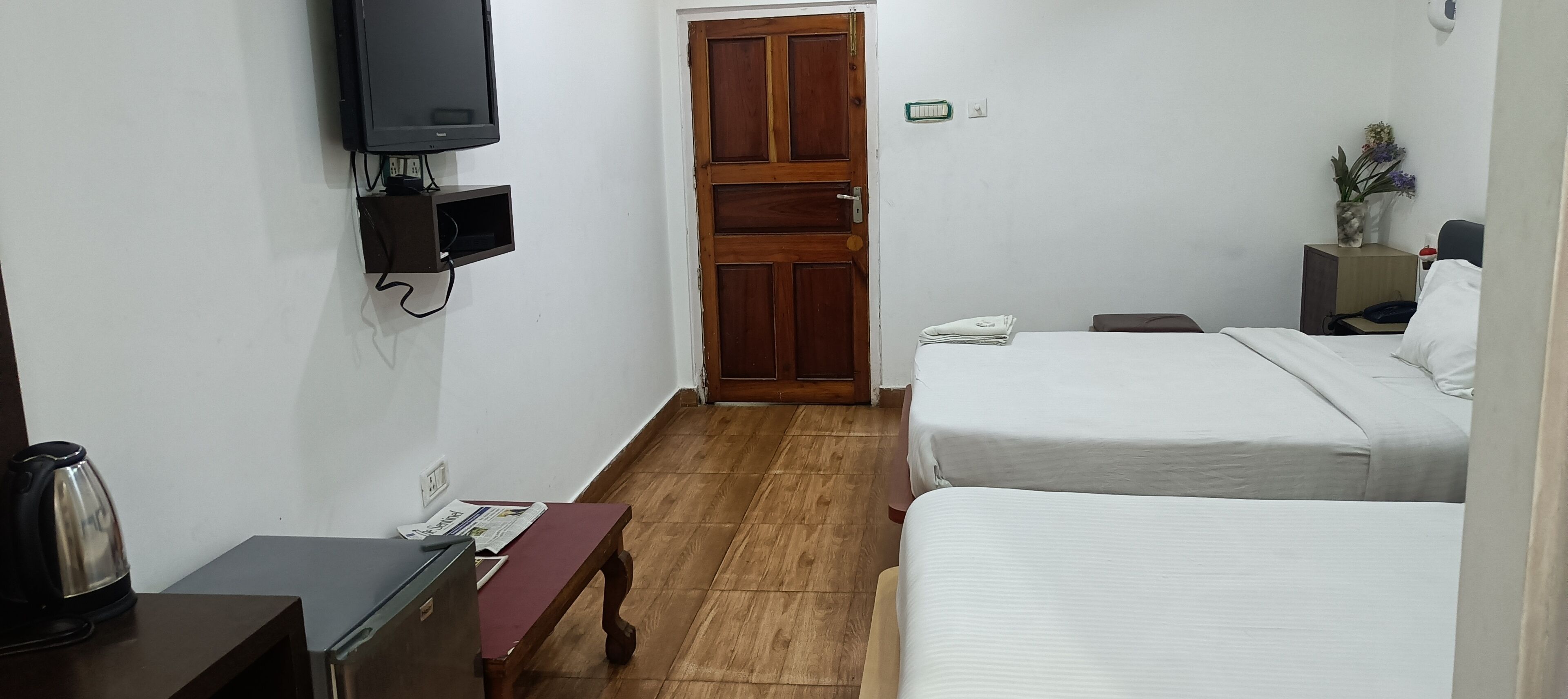 Habitación de hotel con dos camas, un televisor montado en la pared, una puerta de madera, una mesita con una tetera y una mesita de noche con un teléfono y un florero.