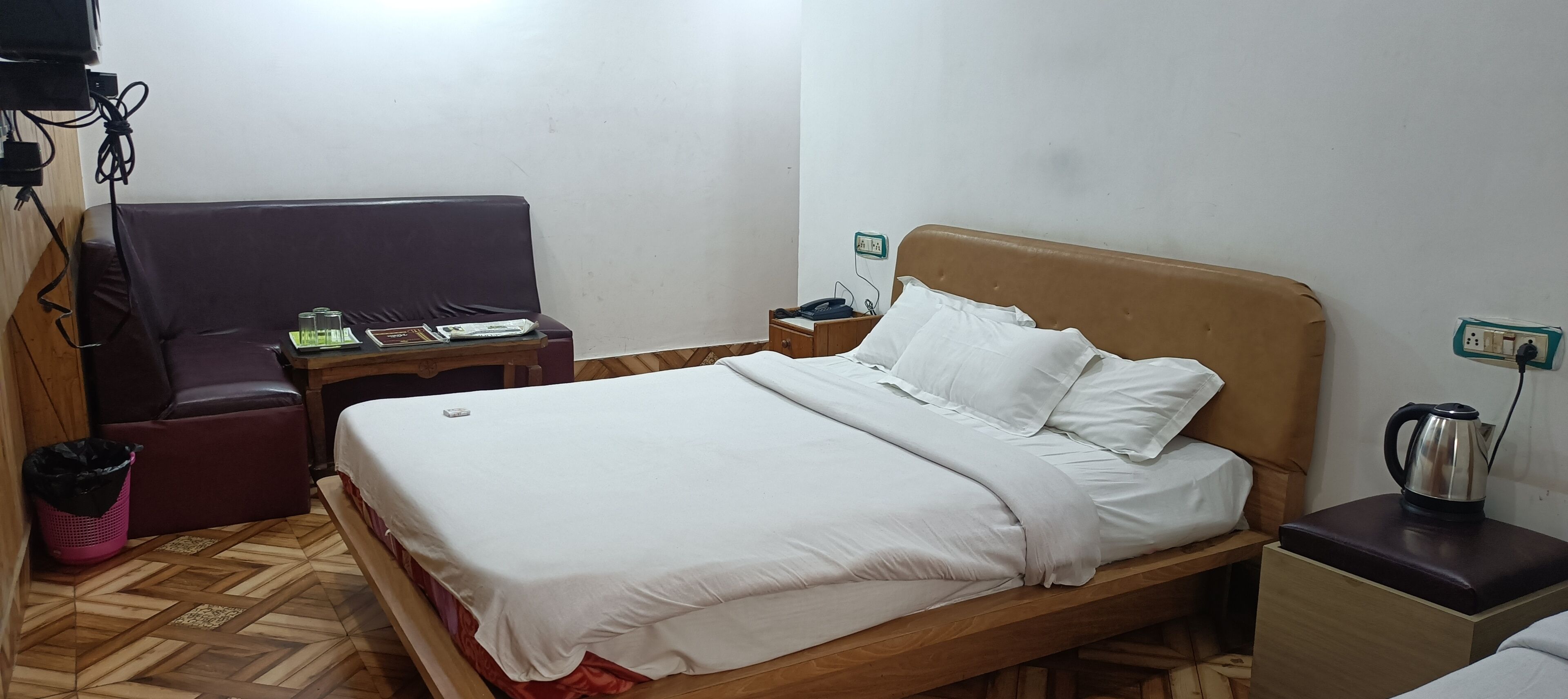 Una cama bien hecha con ropa de cama blanca, un cabecero de madera, una mesita de noche con hervidor y una pequeña mesa con lámpara y libros.