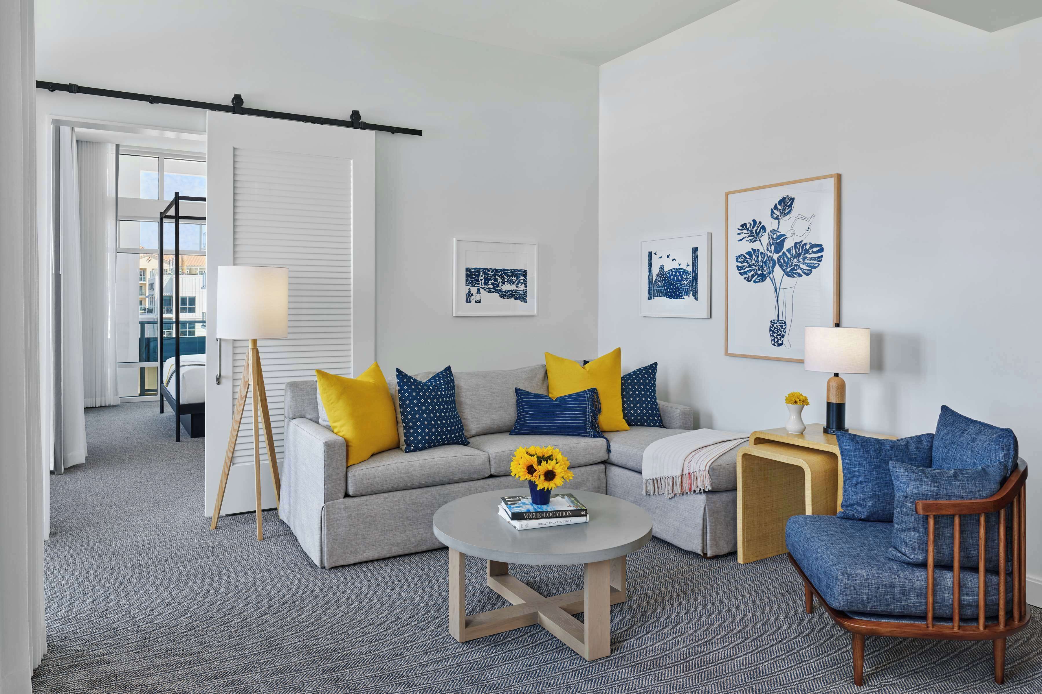 A modern living room with a grey sofa, a round coffee table, and a wooden armchair. There are two side tables with lamps, a bookshelf, and a large window with curtains.