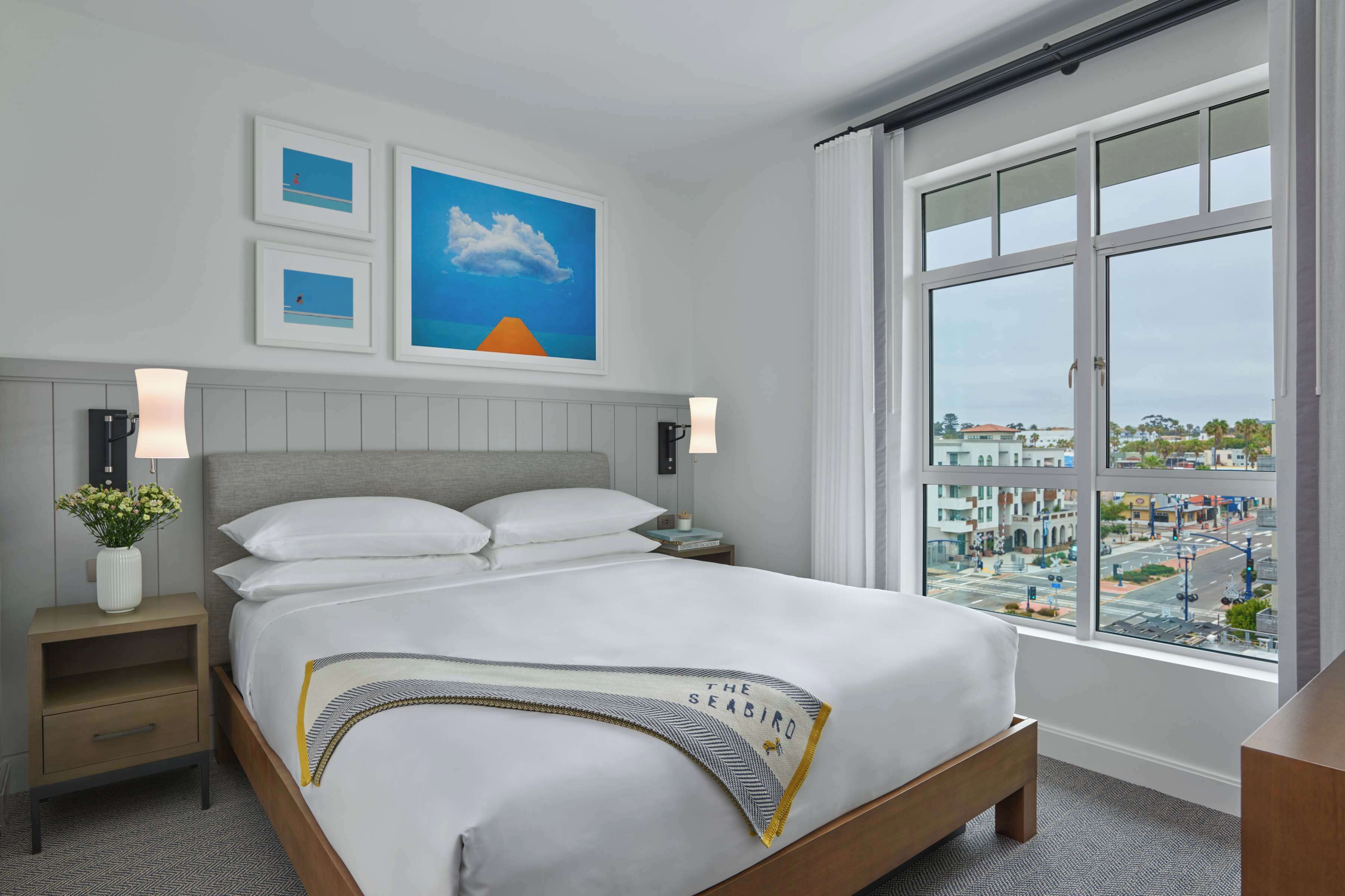 Habitación de hotel con una cama grande, dos mesitas de noche, una ventana con vistas a la ciudad y cuadros en la pared.