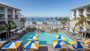 A resort pool area with multiple lounge chairs and umbrellas, a clear view of the ocean, and a building with balconies. - The Seabird Ocean Resort & Spa, Part of Destination Hotel by Hyatt (Oceanside)