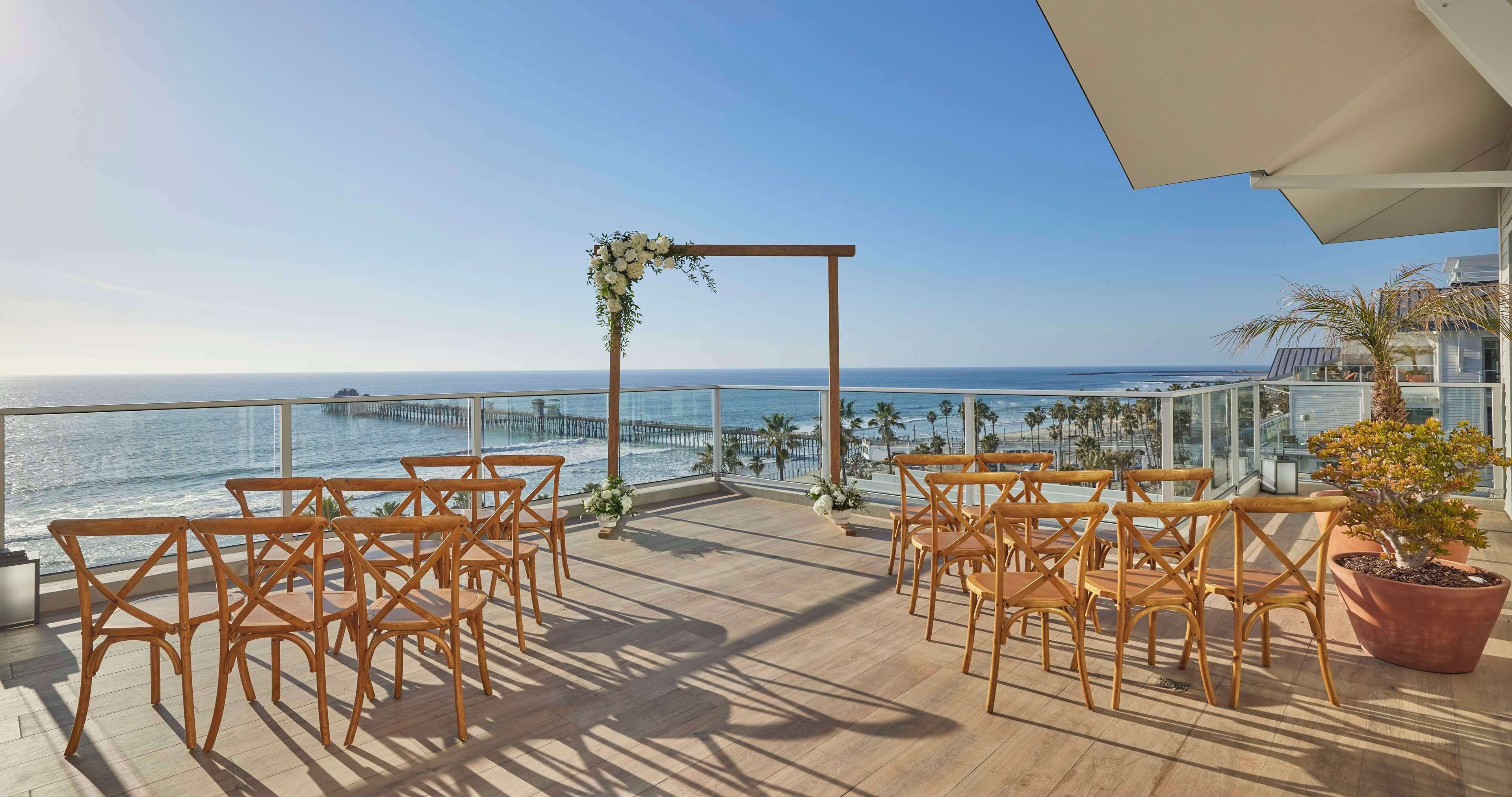 Un balcon agrémenté de chaises en bois et d’une arche ornée de fleurs, offrant une vue sur une plage et l’océan.