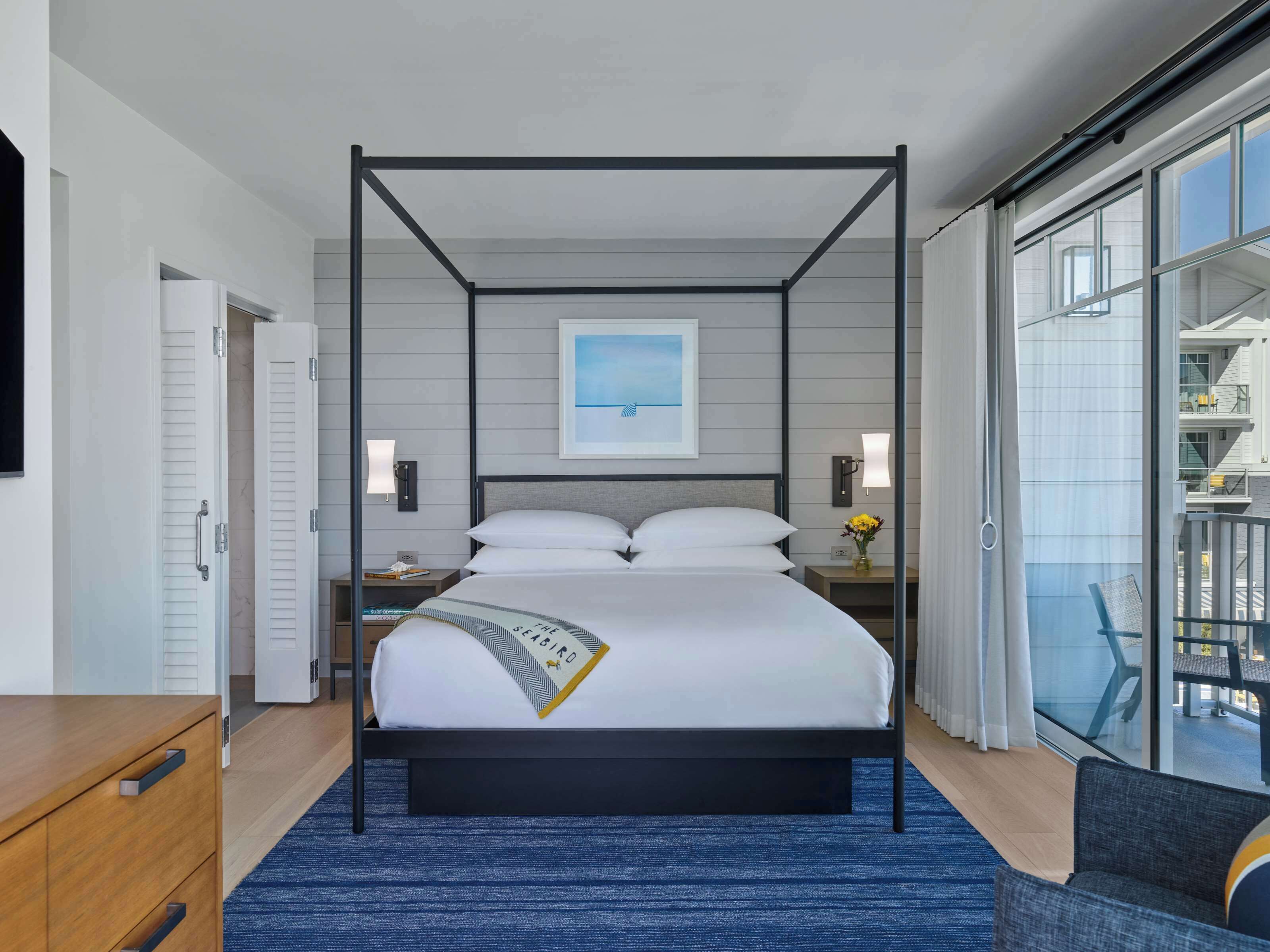 A modern hotel room with a four-poster bed, a large window with a view of the city, a small bedside table with a lamp, and a nightstand with a vase of flowers.