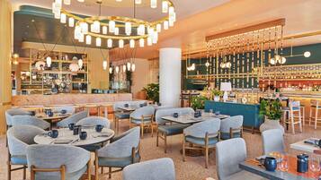Um restaurante moderno com mesas redondas, cadeiras estofadas cinzentas e um bar com prateleiras cheias de garrafas.