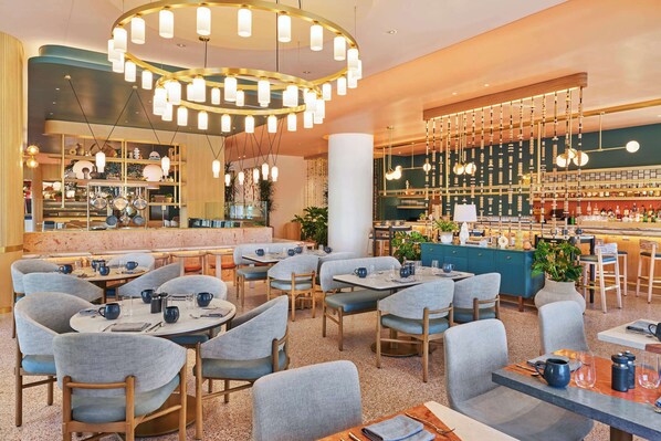 Un restaurante moderno con mesas redondas, sillas tapizadas de color gris y una zona de bar con estantes llenos de botellas.