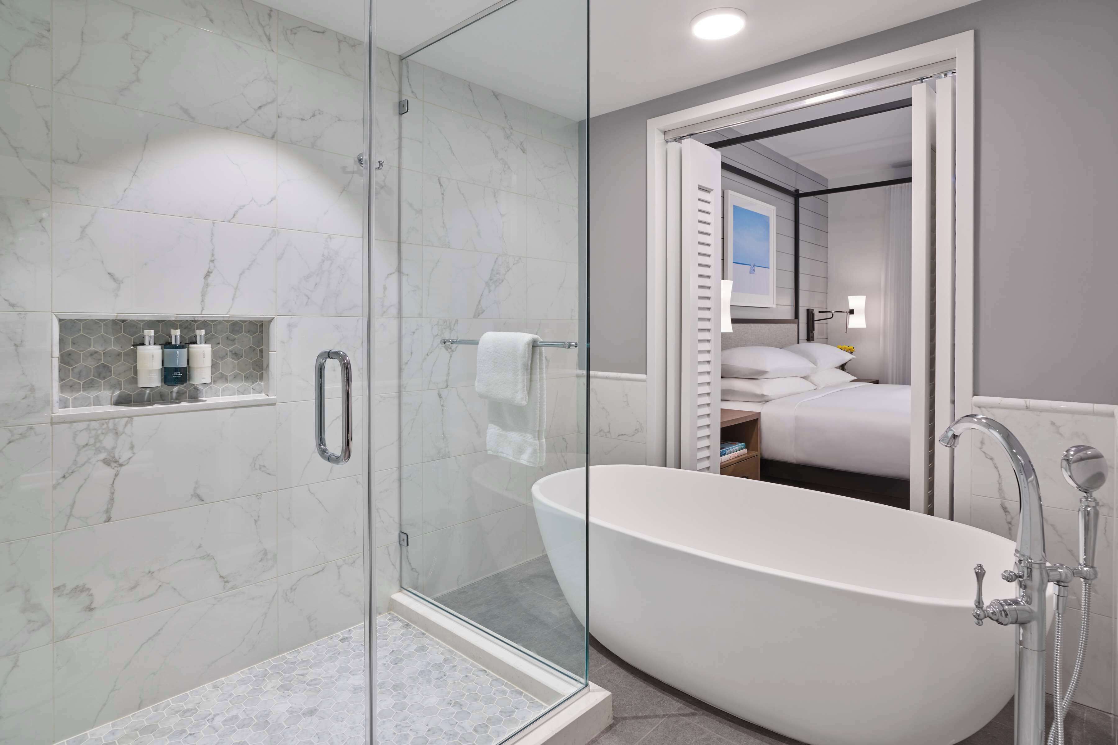 Une salle de bain moderne dotée d’une douche à parois de verre, d’une baignoire sur pieds et d’une chambre visible avec un lit et une fenêtre.