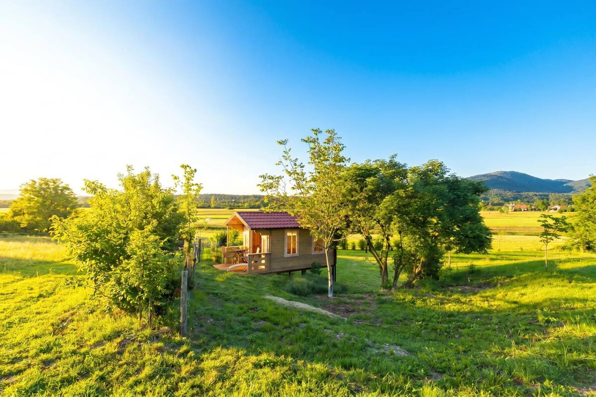A small wooden cabin with a red roof, surrounded by lush green fields and trees, with a clear sky above.