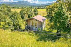 A small wooden cabin with a red roof, surrounded by lush greenery and a fenced area.