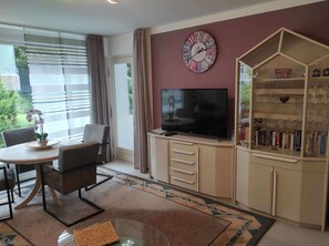 A modern living room with a round table, comfortable seating, a flat-screen TV, a decorative clock, and a glass cabinet with books and decorative items.