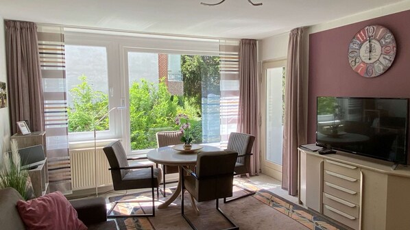 A modern living room with a dining area, large windows, a flat-screen TV, and a decorative clock on the wall.