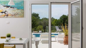 A room with a dining table, chairs, a potted plant, a painting, and a sliding glass door leading to an outdoor pool area. - Villa Deborah (Raša)