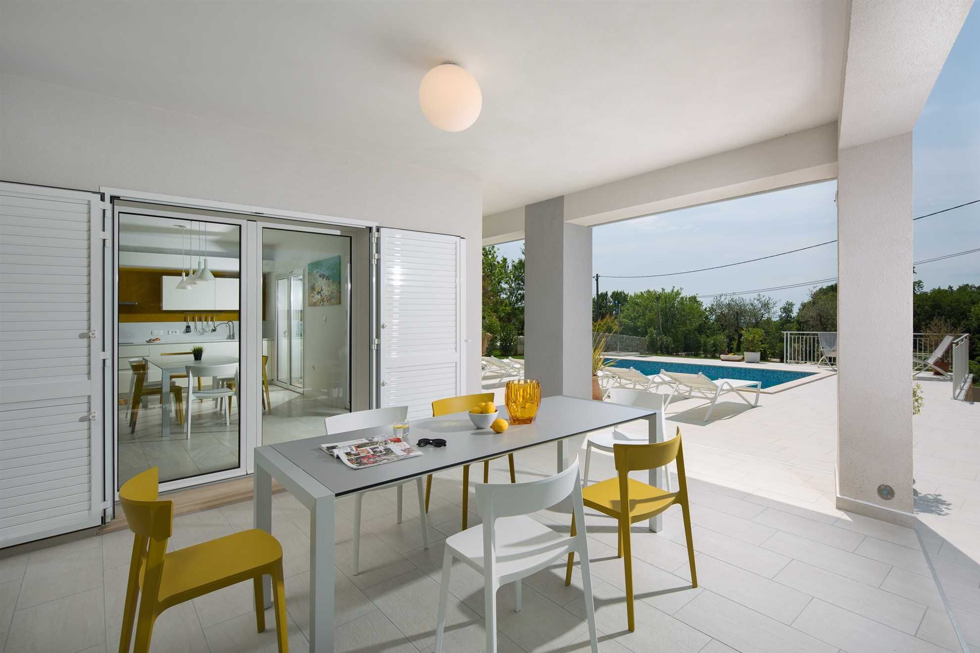 A modern outdoor dining area with a glass table, white and yellow chairs, a pool in the background, and a clear view of the surrounding landscape.