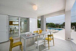 Un espace repas extérieur moderne, avec une table en verre, des chaises blanches et jaunes, une piscine en arrière-plan et une vue dégagée sur le paysage environnant.