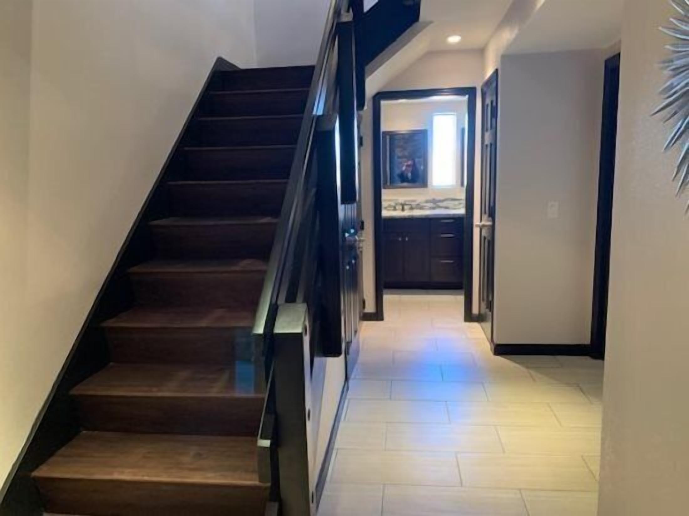 Een moderne trap met glazen balustrade die leidt naar een keuken met donkere kasten en een lichtgekleurde vloer.