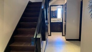 A modern staircase with a glass railing leading to a kitchen area with dark cabinets and a light-colored floor.