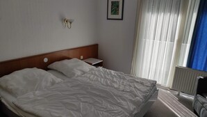 A hotel room with a large bed, a bedside table, a wall-mounted light, a framed picture, and a window with curtains.