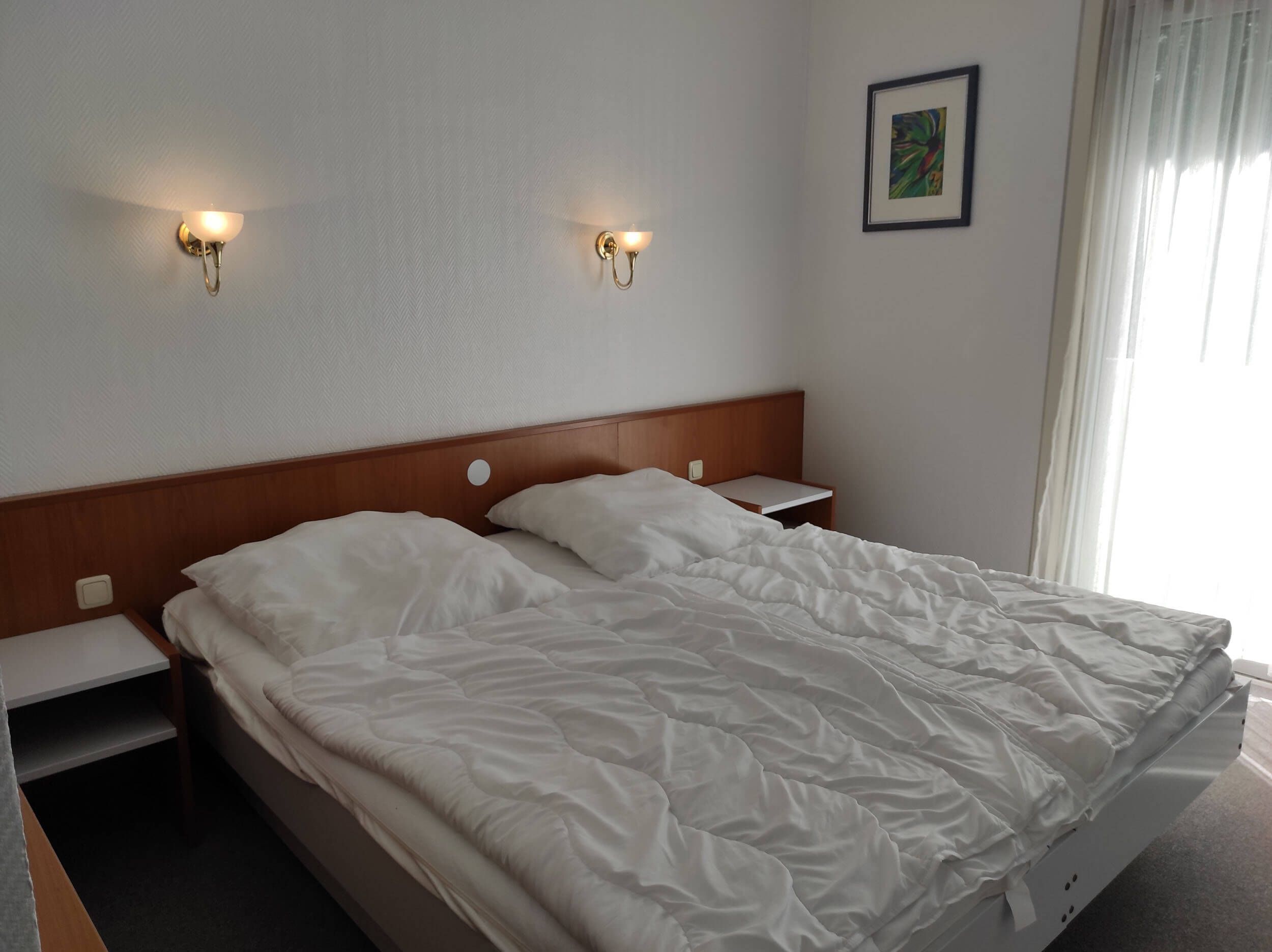 Habitación de hotel con cabecera de madera, dos mesitas de noche, una colcha blanca y dos lámparas fijadas a la pared.