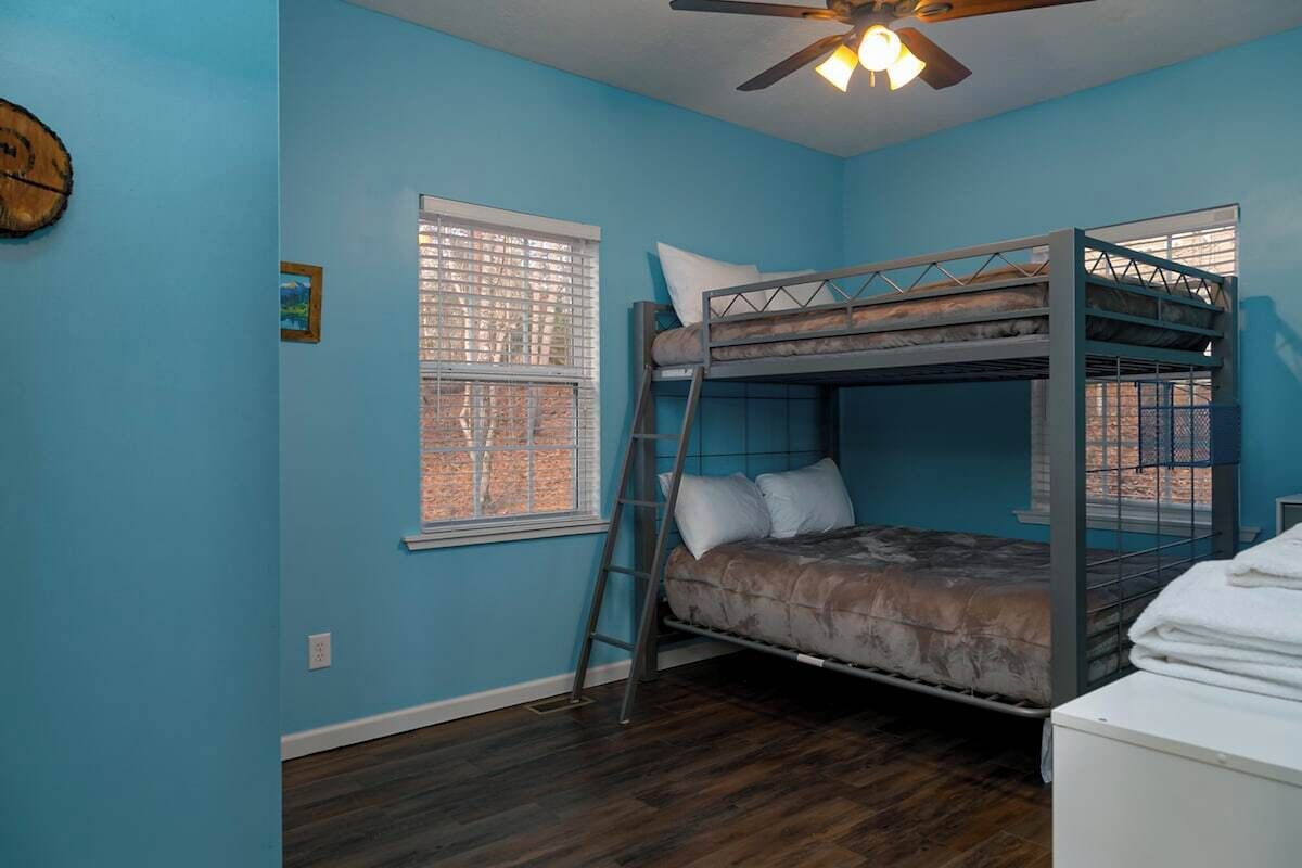A bunk bed room with a ceiling fan, a window with blinds, and a wooden floor.