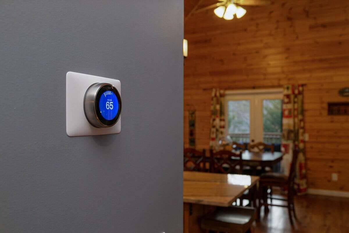 A thermostat displaying 65 degrees, a dining area with wooden furniture, and a window with curtains.