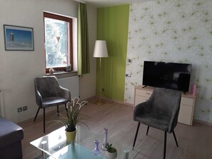 A living room with two chairs, a TV, a lamp, a window with curtains, and a plant on a glass table.