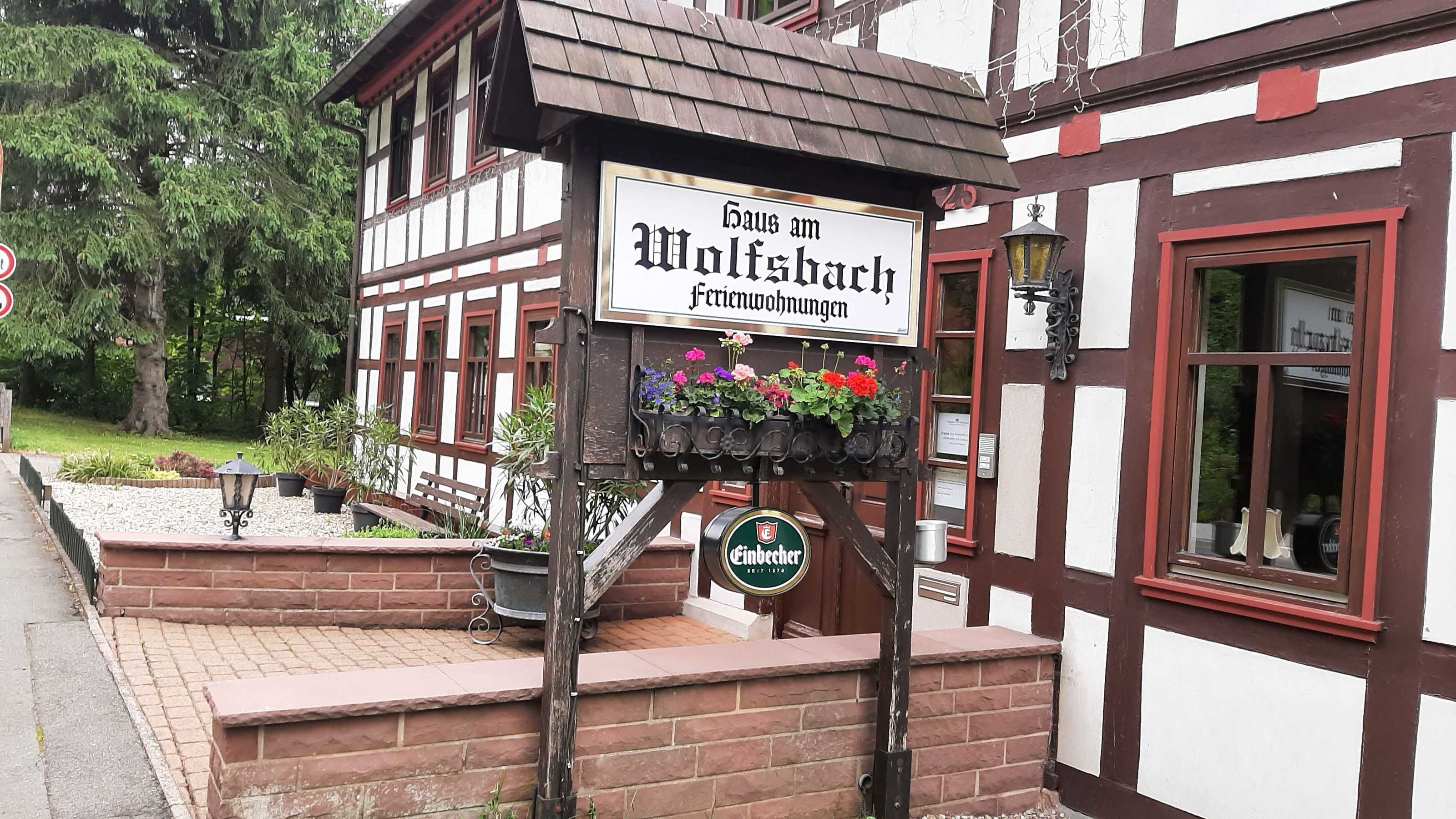 Ein traditionelles Fachwerkhäuschen mit einem Schild, das 'Haus am Wolfsbach Ferienwohnungen' und ein Bierzeichen darunter zeigt.