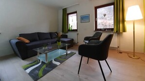 En stue med en sofa, et sofabord i glas, en lænestol, en lampe og udsigt til et sneklædt landskab gennem vinduerne.