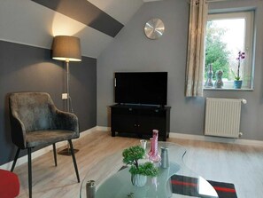 A living room with a flat-screen TV, a lamp, a chair, a glass coffee table with decorative items, a clock, and a window with a view of greenery. - Staufenberg Vacation Rental - House in Wolfsbach - Vacation Apartments (Walkenried)