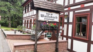 En traditionell byggnad med korsvirkesfasad, med en skylt som visar texten ”Haus am Wolfsbach Ferienwohnungen” och en ölreklamskylt under.