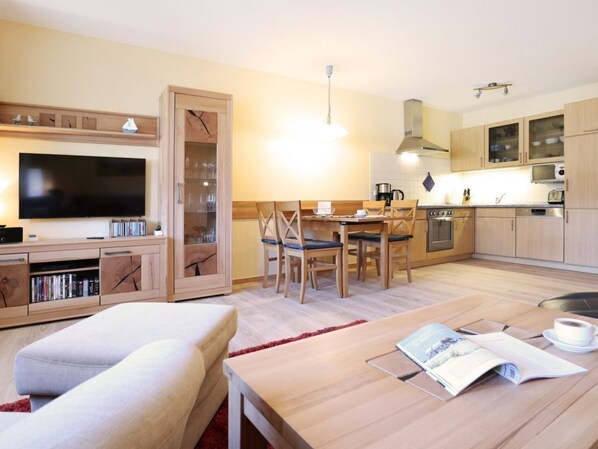 A modern living room with a flat-screen TV, a sofa, a dining table with chairs, and a kitchen area with cabinets and appliances.