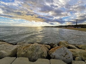 A coastal scene with a rocky shoreline, a beach, and a distant view of a cityscape under a cloudy sky.