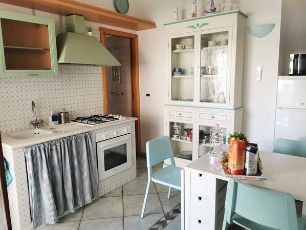 A compact kitchen with a gas stove, sink, and oven. There is a dining area with a table and chairs. A cabinet with glass doors is visible, containing various kitchen items.