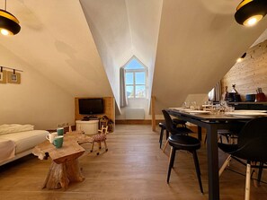A cozy attic bedroom with a sloped ceiling, a television, a wooden dining table, and a view of the outdoors through a window.