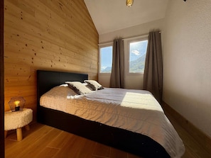 A bedroom with a wooden ceiling, a bed with white bedding, a nightstand with a lamp, and a window with curtains.