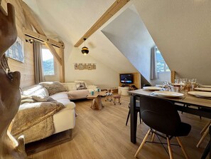A cozy loft-style room with a wooden ceiling, a sofa, a dining table set for four, and a television. - 4-room apartment for 10/12 people in Briançon with Wifi (Briançon)