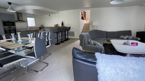 Un salon moderne avec un canapé gris, un tapis blanc et moelleux, et un coin repas comprenant une table en verre et des chaises en métal.