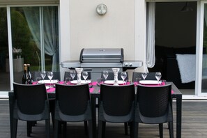 Ein Essbereich im Freien mit Grill, schwarzen Korbstühlen und einem Tisch, der mit rosa Tischsets und Weingläsern gedeckt ist.