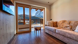 A spacious living room with a wooden floor, a sofa with fur throw pillows, and a large sliding glass door leading to a balcony with a mountain view.