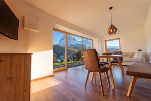Een moderne woonkamer met een groot raam en uitzicht op de bergen, een eethoek met houten meubels, en een flatscreen televisie die aan de muur is bevestigd.