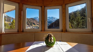 Une pièce avec un plafond en bois, une table ronde recouverte d’une nappe blanche et trois fenêtres donnant sur des montagnes et des forêts.