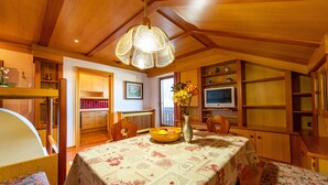 Un espace de repas accueillant et bien éclairé, avec un plafond en bois, une table dressée avec un saladier de fruits et un vase, et un téléviseur posé sur une étagère.
