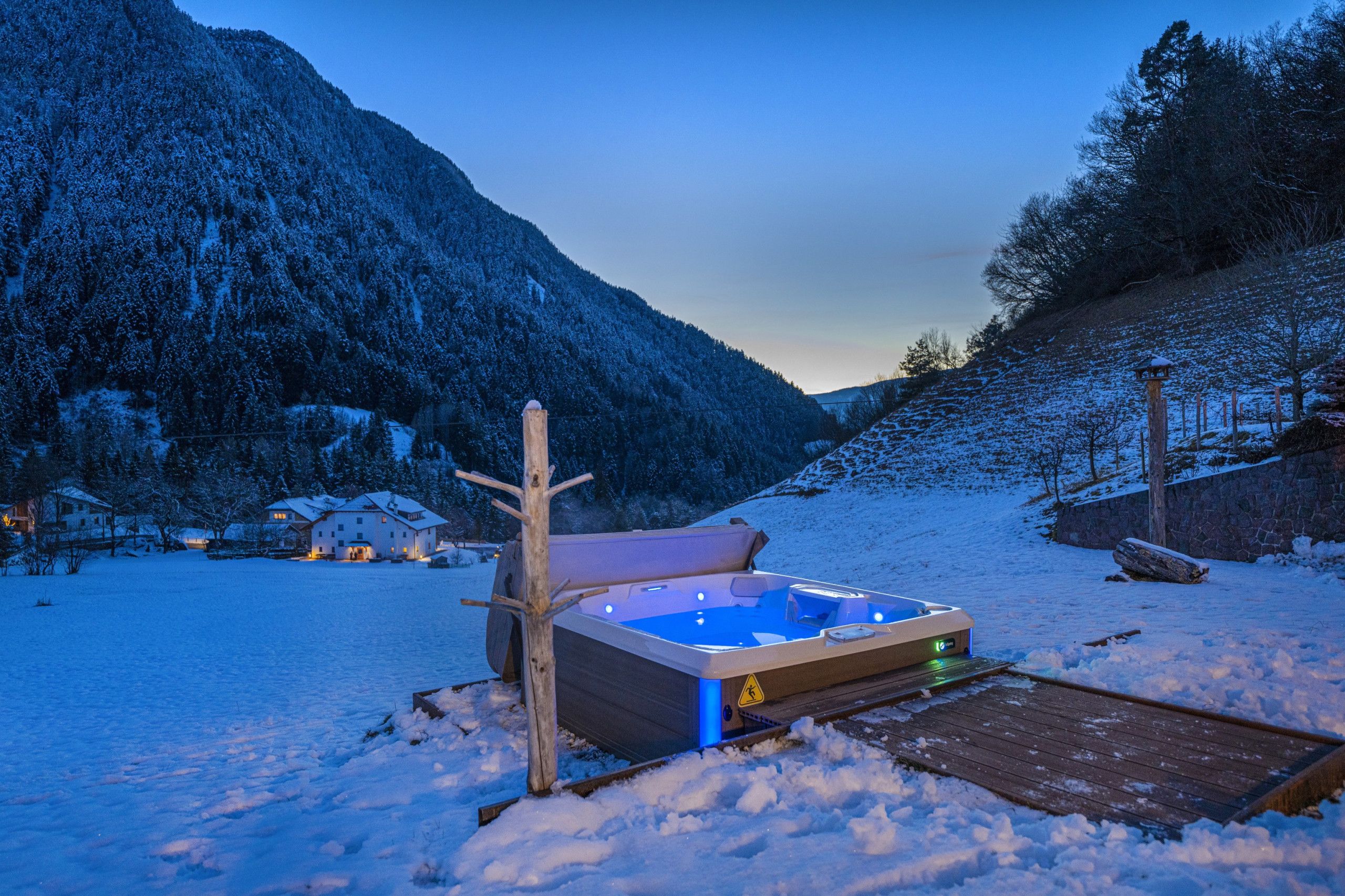 Un paisaje nevado con una terraza de madera que conduce a un área de hidromasaje, rodeado de árboles cubiertos de nieve y una montaña al fondo.