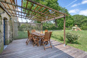 Une terrasse en bois avec une pergola, une table et des chaises, et une vue sur une aire de jeux en arrière-plan.