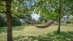 Een hangmat tussen twee bomen in een gazon, met huizen op de achtergrond.